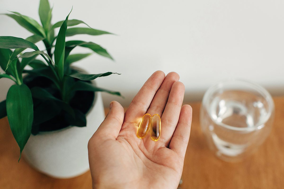 The photo is focused on a woman's hand which is holding two Omega-3 capsules. There is a glass of water and a houseplant in the background.
